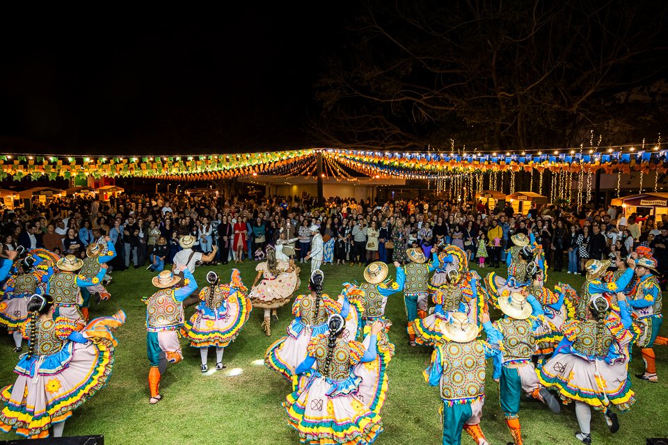 Quadrilha junina se apresenta ao ar livre para uma grande plateia, sob decoração de bandeirinhas coloridas, durante o Arraiá do Brasília Palace.