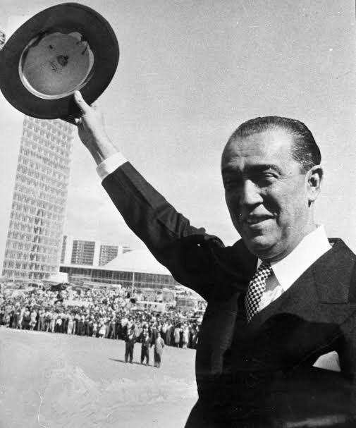 Juscelino Kubitschek acena para a multidão durante a inauguração de Brasília, com o edifício do Congresso ao fundo.